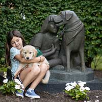 Little girl holding a small puppy right next to a statue of a girl with a seeing eye dog